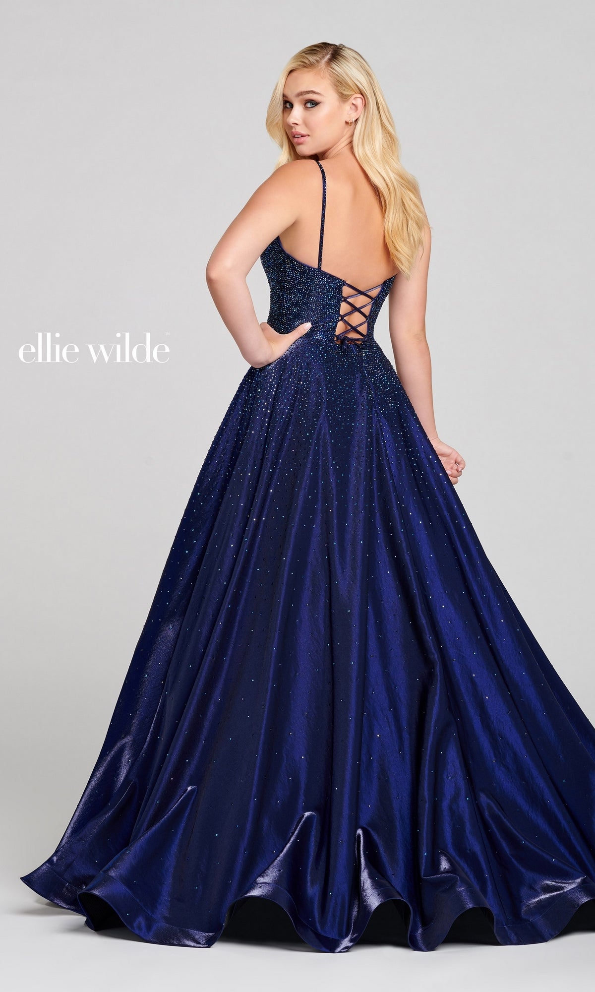 A woman poses against a plain background in the Ellie Wilde Ew121005 long beaded prom ball gown, featuring deep blue sparkles, an A-line silhouette, and a lace-up open back.